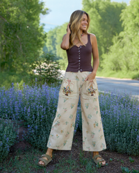 A woman standing outdoors in a field of purple flowers, wearing a dark brown sleeveless button-front top and cream wide-leg pants with a floral print and embroidered pocket details.