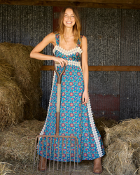 A woman stands in a barn holding a pitchfork, wearing a sleeveless boho-style maxi dress with a blue, pink, and white floral pattern and white lace trim.