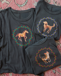 Three dark charcoal gray crewneck t-shirts laid flat on a patterned rug, each featuring a different animal illustration inside a floral wreath: a horse on the largest shirt, a dog on the middle shirt, and a cat on the smallest shirt.