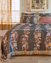 A bed dressed in a dark charcoal chocolate floral blanket and matching pillow shams, styled in a warm, rustic bedroom with wood floors, a window with sheer curtains, and a wooden wall cabinet.