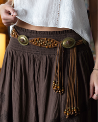 A close-up of a person wearing a brown skirt with a decorative woven waist belt featuring large oval metal conchos and dangling beaded cords, paired with a white cropped top.