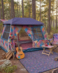 A colorful camping tent set up in a forest with a matching patterned floor mat, folding chair, small folding table with a lantern, and an acoustic guitar leaning against the tent.