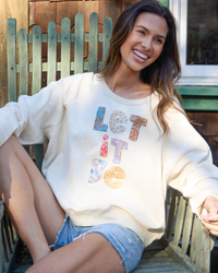 Woman sitting in chair wearing cream sweatshirt with mix printed letters that say 'Let It Be'