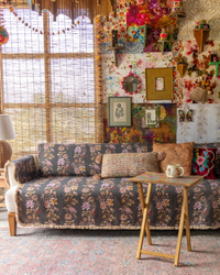A bohemian-style living room featuring a dark gray floral-patterned sofa, a small wooden side table with a teacup, and a highly decorated wall filled with colorful framed art, tapestries, and decorative accessories.