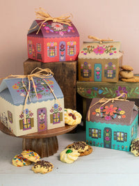 Decorative house-shaped gift boxes with colorful designs on a wooden surface with cookies.