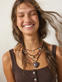 A smiling woman with long brown hair wearing a layered bohemian necklace with colorful beads and a dark heart-shaped pendant, paired with a brown ribbed tank top against a plain light background.