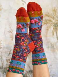 A pair of crew socks with a colorful floral and geometric pattern, worn on feet against a soft beige background with dried grass silhouettes.